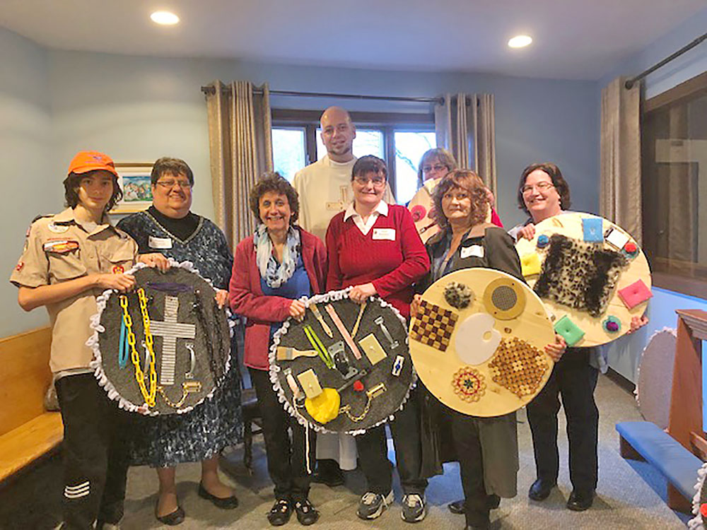 Drew Newberry, the Open Door Committee (Sharon Urbaniak, Heidi Mack, Patty Wittman, Judy Lake and Mary Regula, and Paula Potteiger(back row))  with Father Bryan Zielenieski after the Inclusive Easter Mass holding the finished sensory boards. 
Courtesy of Sharron Urbaniak